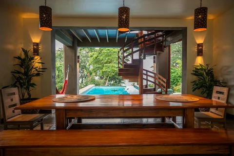 A timber dining table beneath hanging lamps, with the pool and spiral staircase beyond.